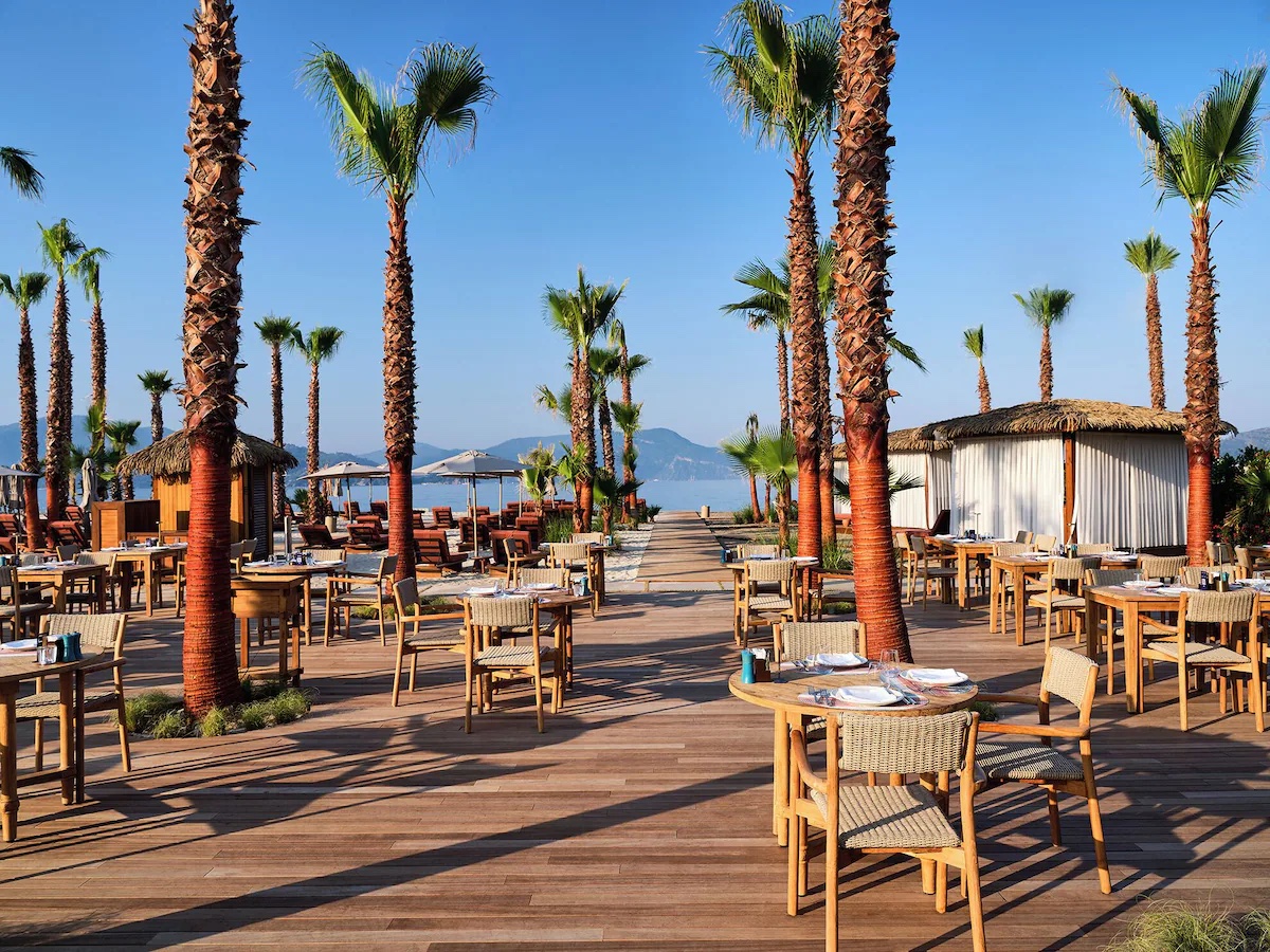 Cape Arna XO Fethiye restoran terasında palmiyeler arasında ahşap masa ve sandalyeler